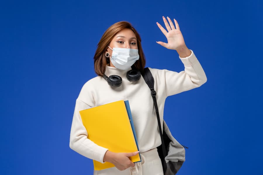 front view young female student white jersey wearing mask backpack holding files waving blue wall (1)